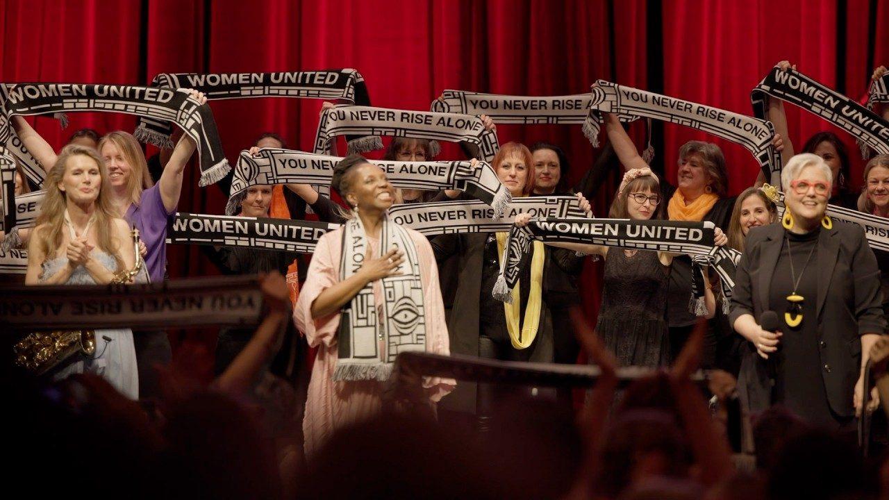 Künstlerinnen auf der Bühne halten Schals mit den Aufschriften „WOMEN UNITED“ und „YOU NEVER RISE ALONE“ hoch